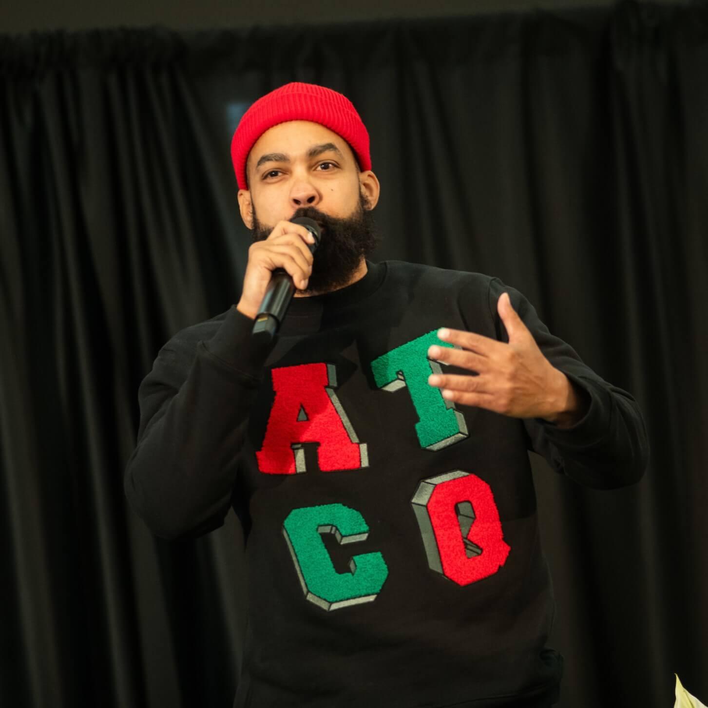 Jasiri X holding a microphone in front of a black backdrop. He is wearing a red cap and a black A Tribe Called Quest crewneck.