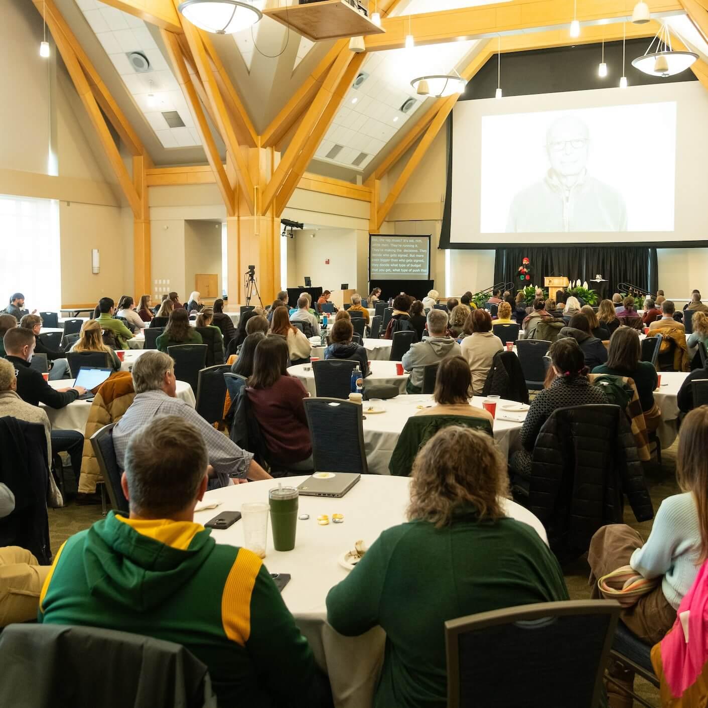 A full house in the Silver Maple ballroom sitting at tables looking at a presenter in the front row. An image of Harry Belafonte is projected on a large screen.