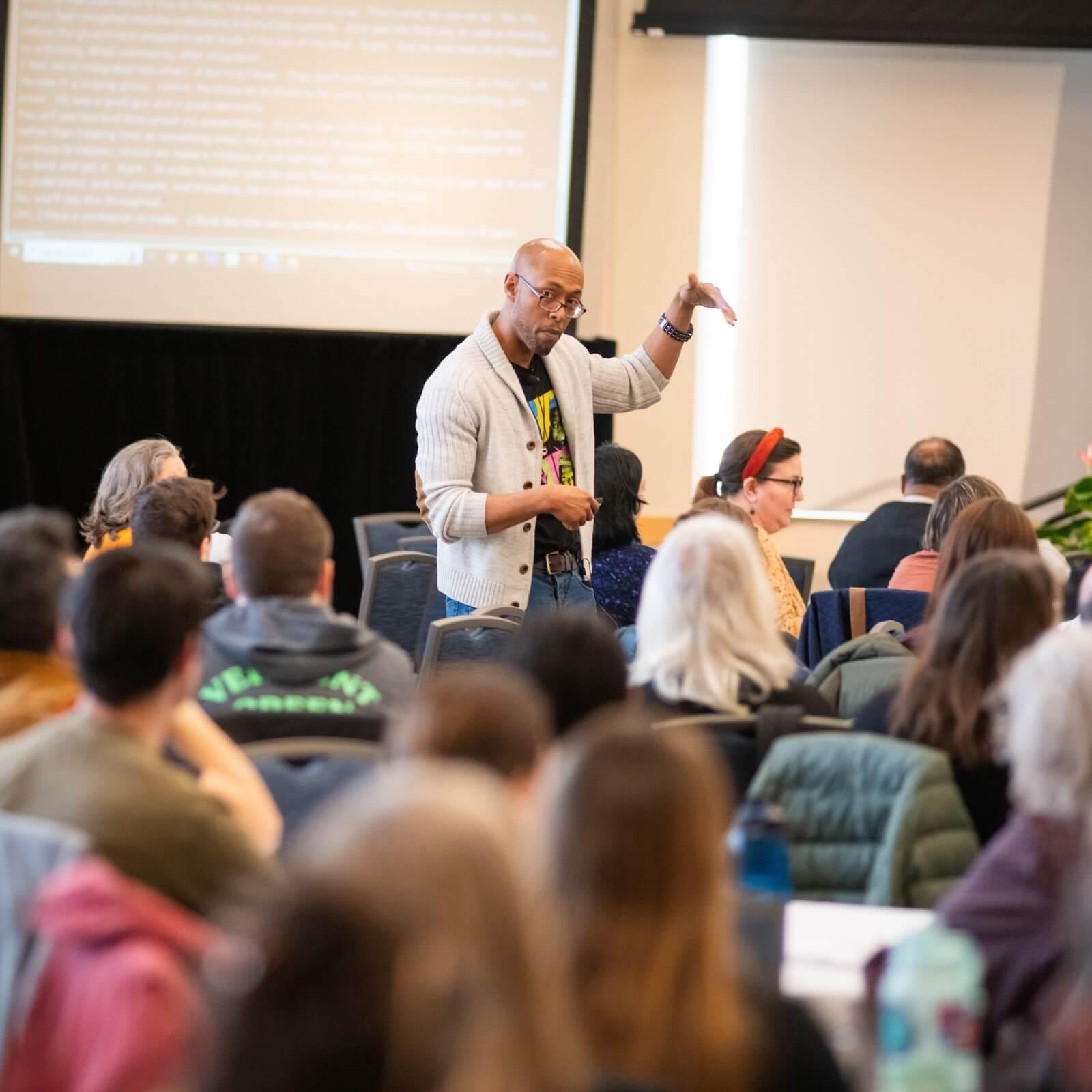 Delma Jackson III standing amongst a seated audience speaking. He is gesturing with one hand held near his head.