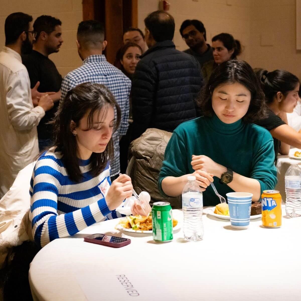 Two young women sitting and eating at a table with a group of people standing and talking behind them.