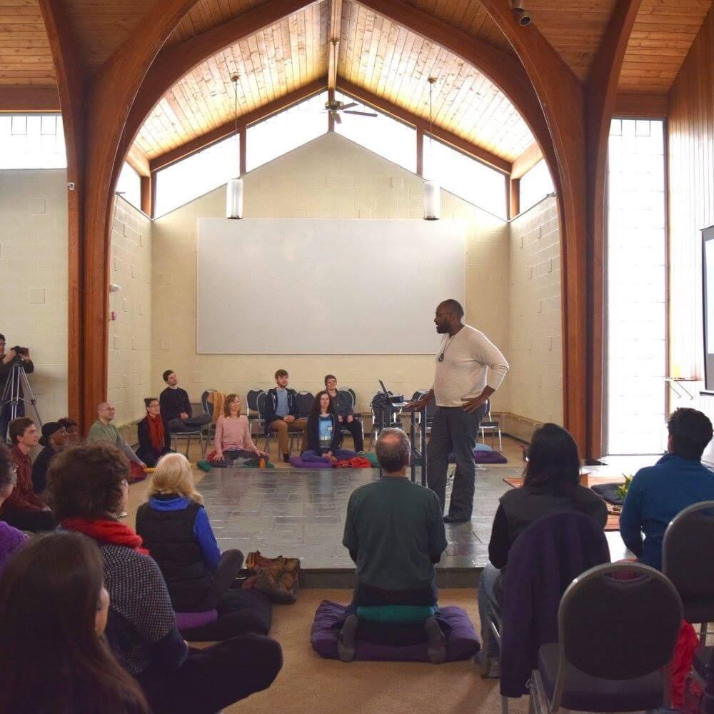 People sit on the floor and in chairs listening to a speaker in the middle of the room on a raised tile floor.