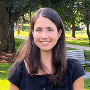 Janine, smiling and standing near a shady tree and wooden bench