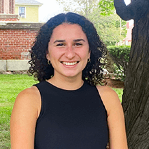 Jill, smiling and standing under a large tree, surrounded by green grass and a brick wall..