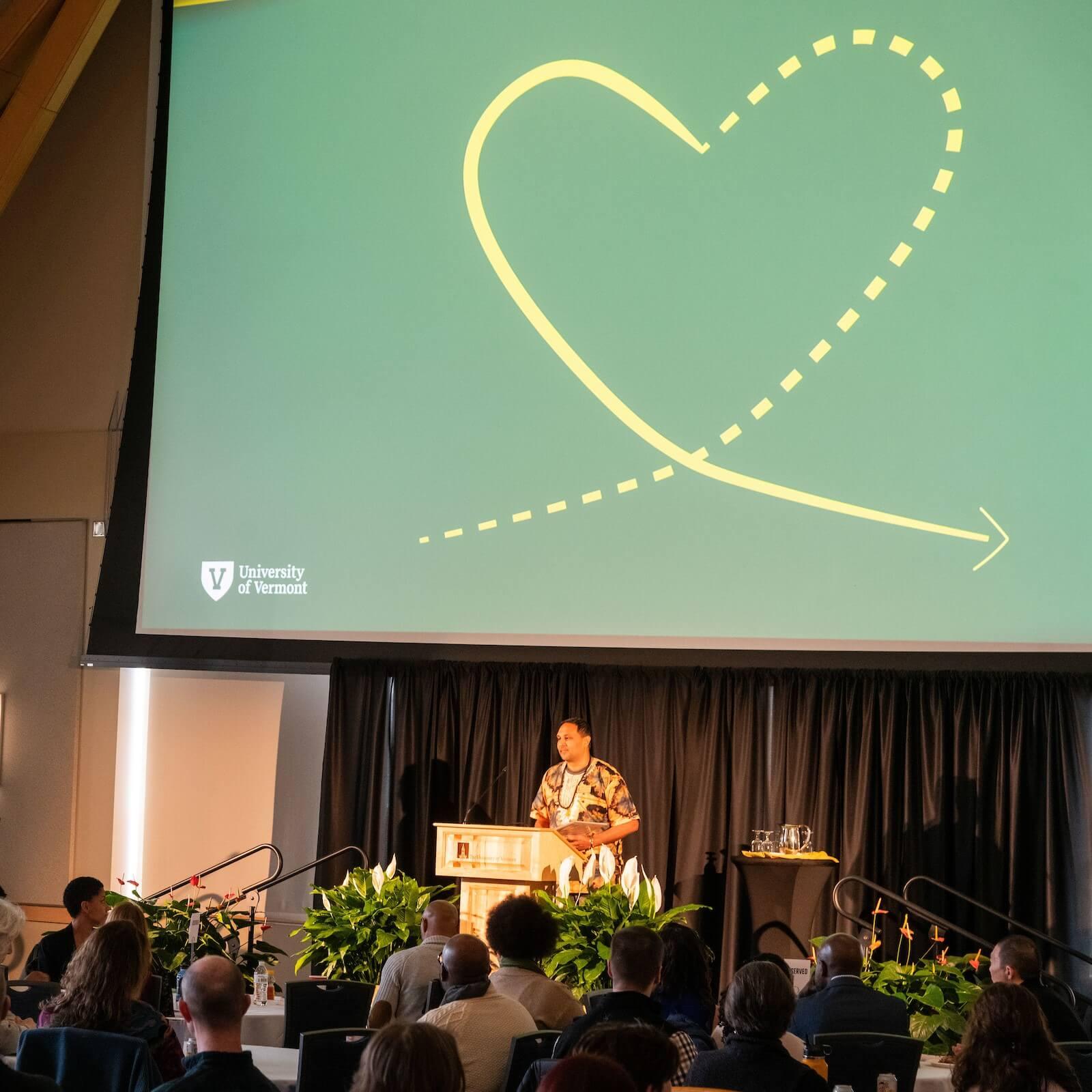 Vice Provost Amer Ahmed speaking at a podium. Behind him is an image of the 2024 IES logo: a dotted line that curves into a solid line heart shape with an arrow at the end of it.