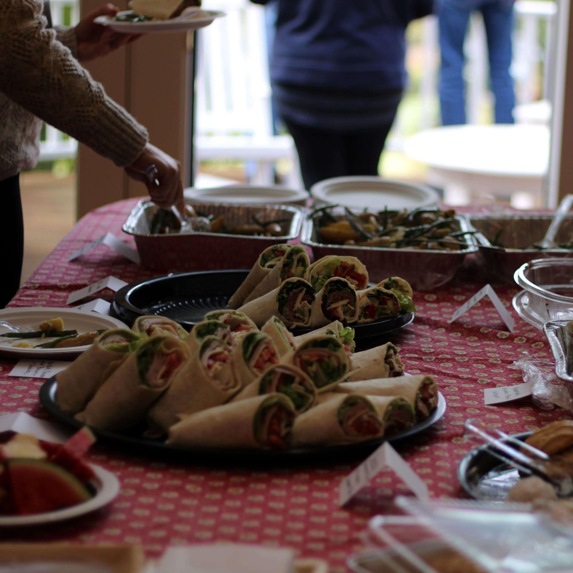 A spread of food on a table cloth with a person's arm holding a utensil grabbing something from a tray.