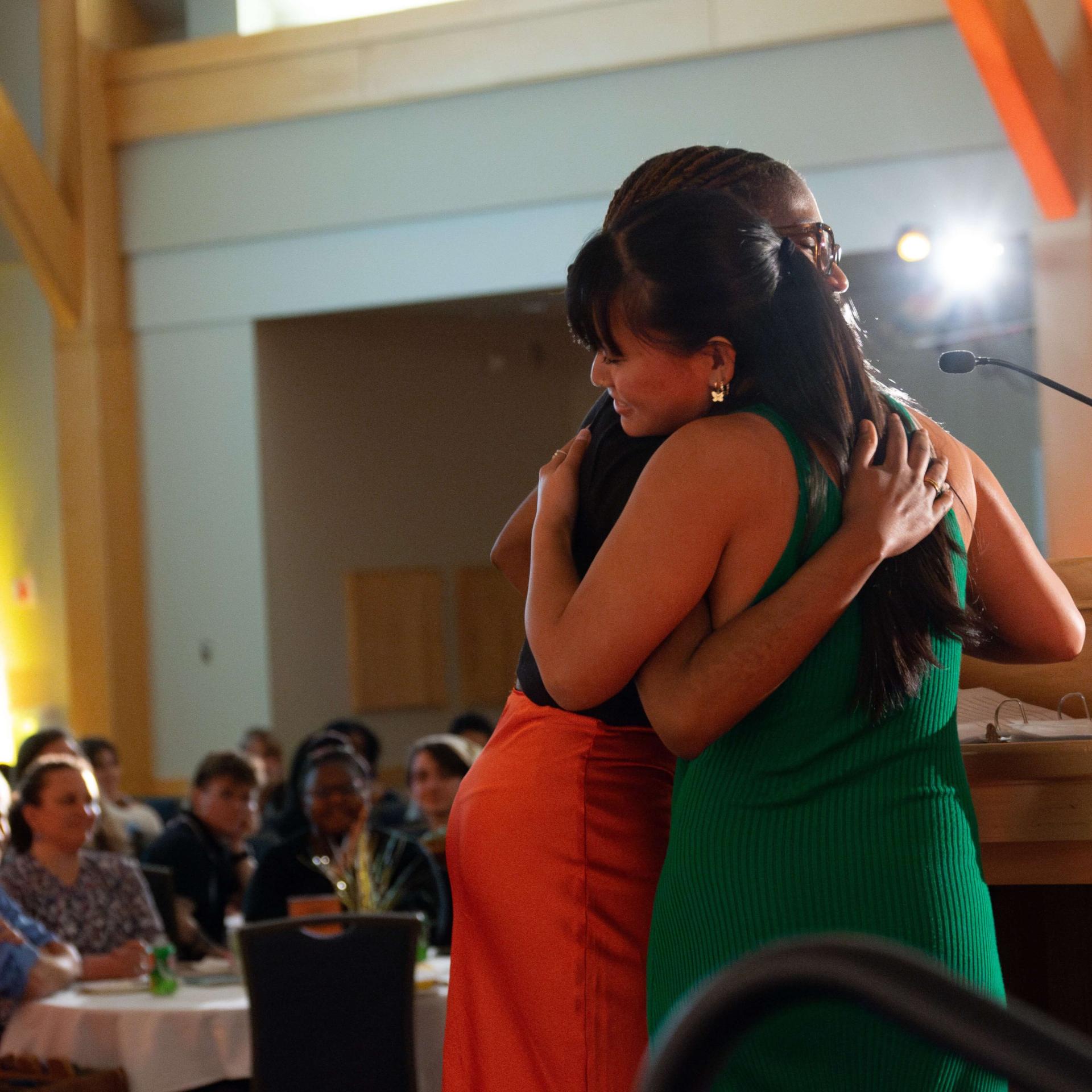 Two young women wearing semiformal outfits hug at a podium in front of a cheering crowd