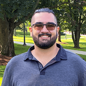 Marc, wearing sunglasses and smiling surrounded by shady trees and green grass.