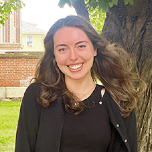 A smiling Mathilde, under a tree near a brick wall.