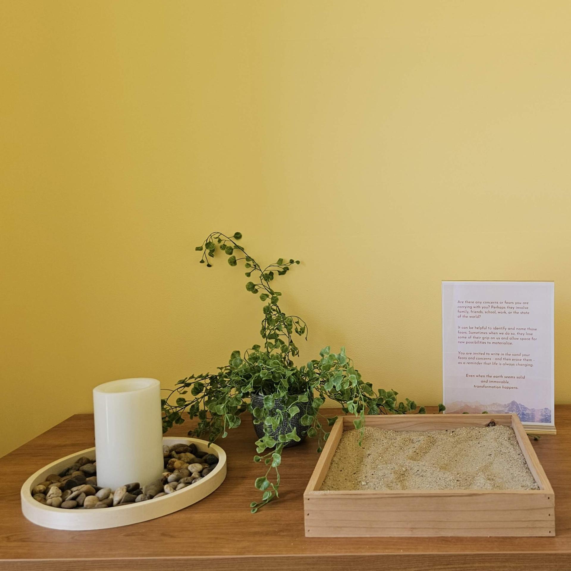 Serene yellow wall with a plant, sand raking kit, and a candle surrounded by pebbles on top of a cabinet.