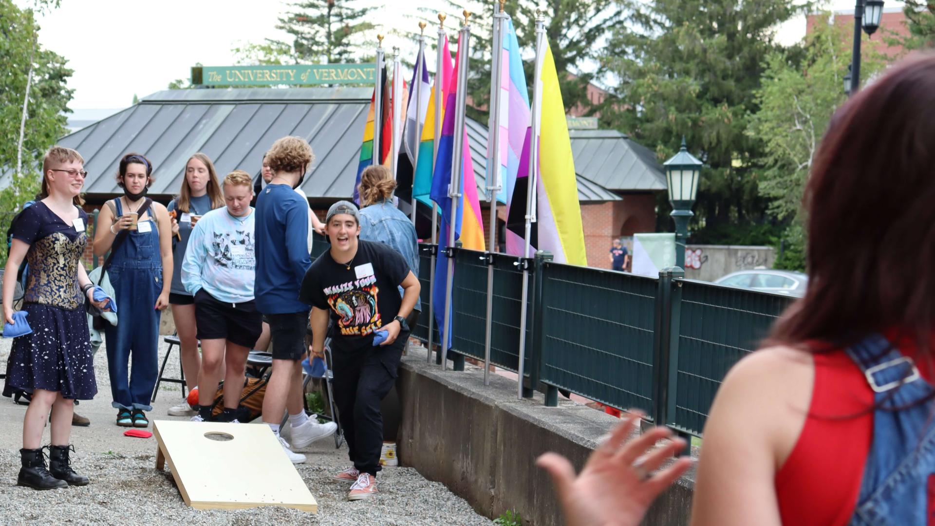 Students play a game of cornhole in front of different LGBTQ+ identity flags on an outdoor patio.