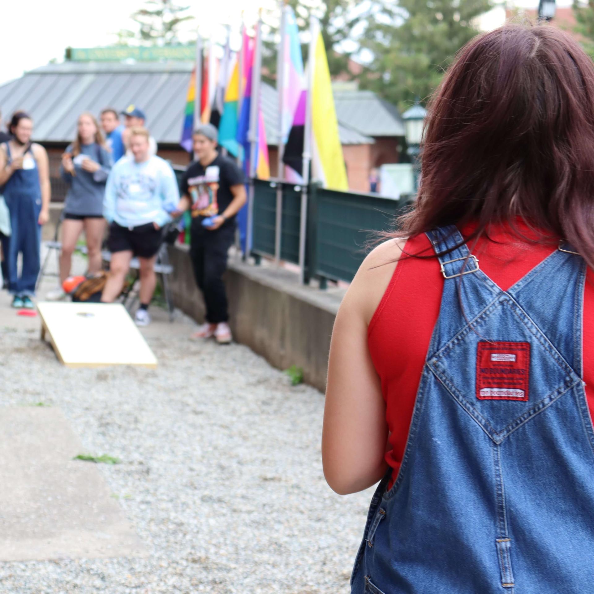 Person with long hair, red tank top, and overalls is about to throw a bean bag in a game of cornhole. In the background is another group of students and different LGBTQ flags.