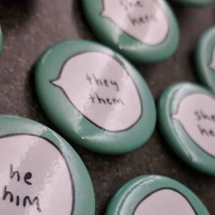 Blue buttons laid out on a table. Each button has a word bubble with different pronouns in them, including he/him, they/them, she/her.