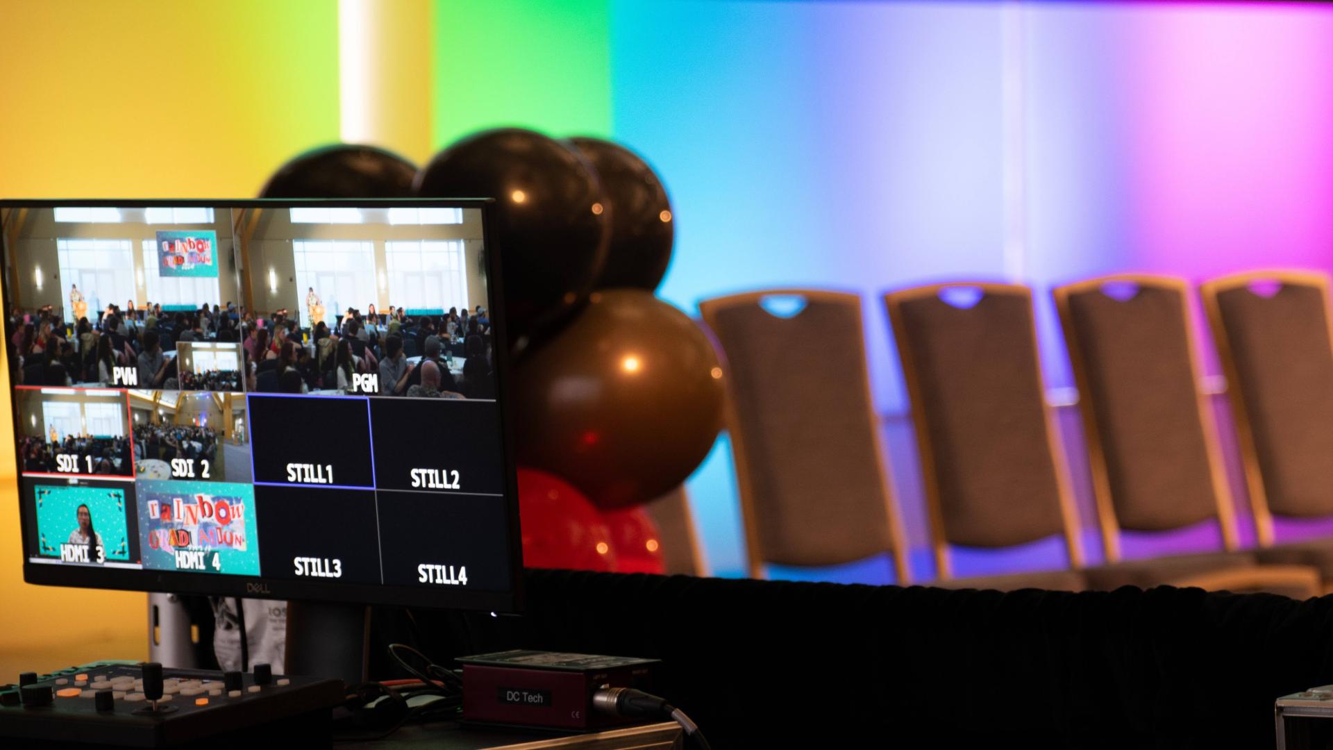 Tech screen showing multiple video inputs, including a person on zoom, an audience, and a slide that says "Rainbow Graduation" all in front of colorful balloons, a row of empty chairs, and rainbow uplighting against a back wall.