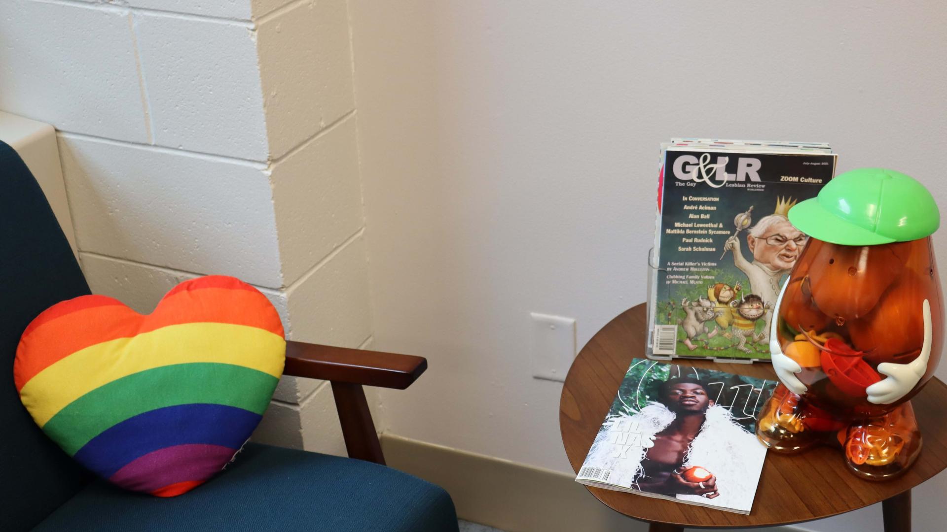 A chair with a rainbow heart pillow on it next to a small end table that holds copies of queer magazines and a Potato Head-shaped jar containing more Potato Heads inside.