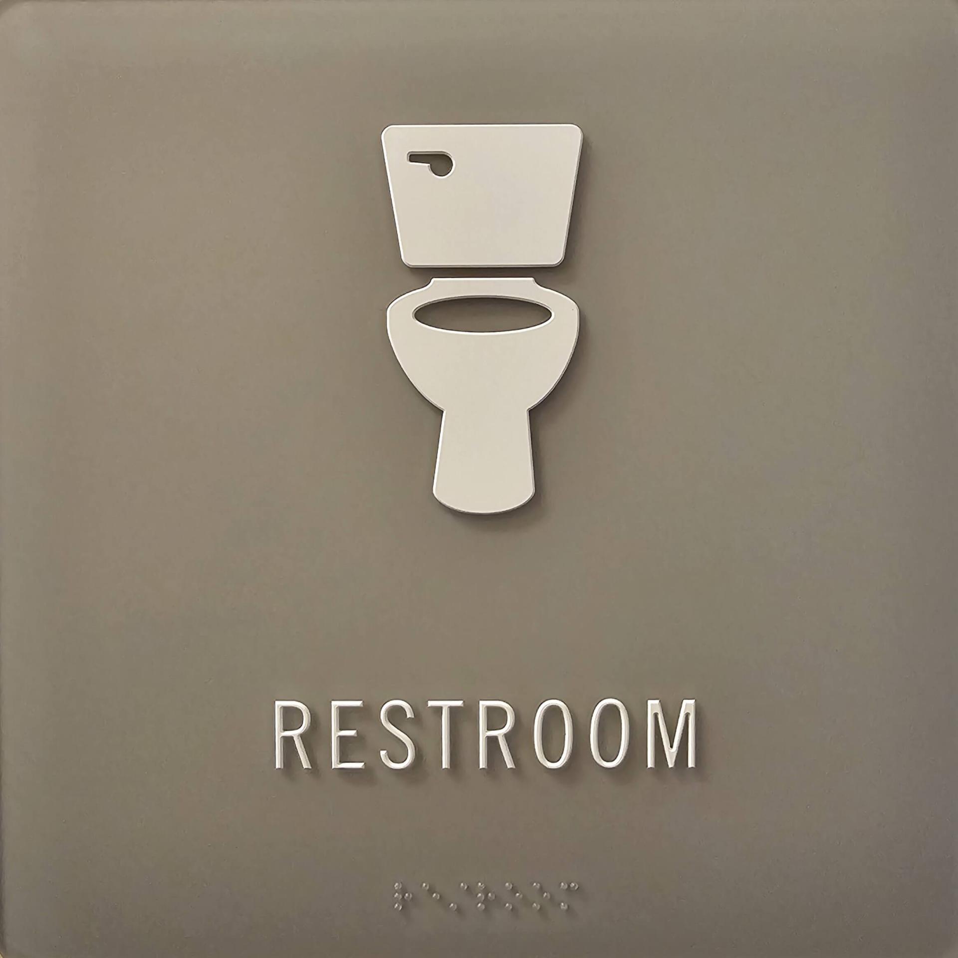 A grey restroom sign with a icon of a toilet, the word "restroom" written in English and embossed in braille. 