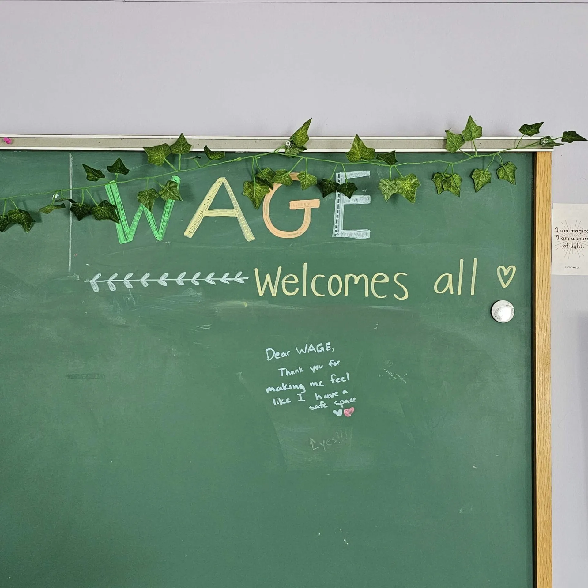the corner of green chalkboard with garland around the top against a pale purple wall with the message, "WAGE welcomes all!" written on it.
