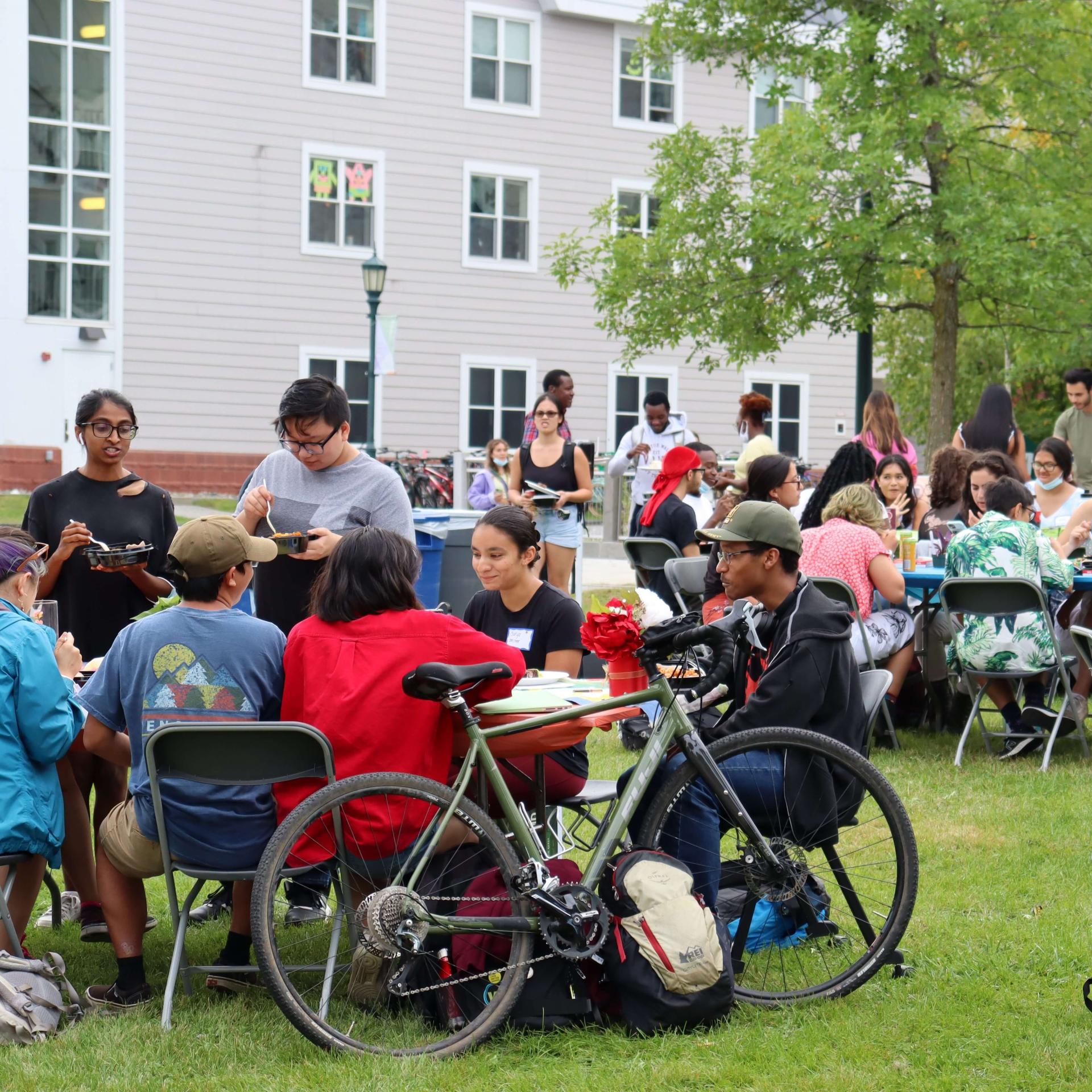 Students gather on a green sitting in chairs at a few folding tables, eating and talking.