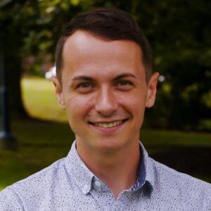 Brandon smiling and out on a green space with trees, shrubs and grass