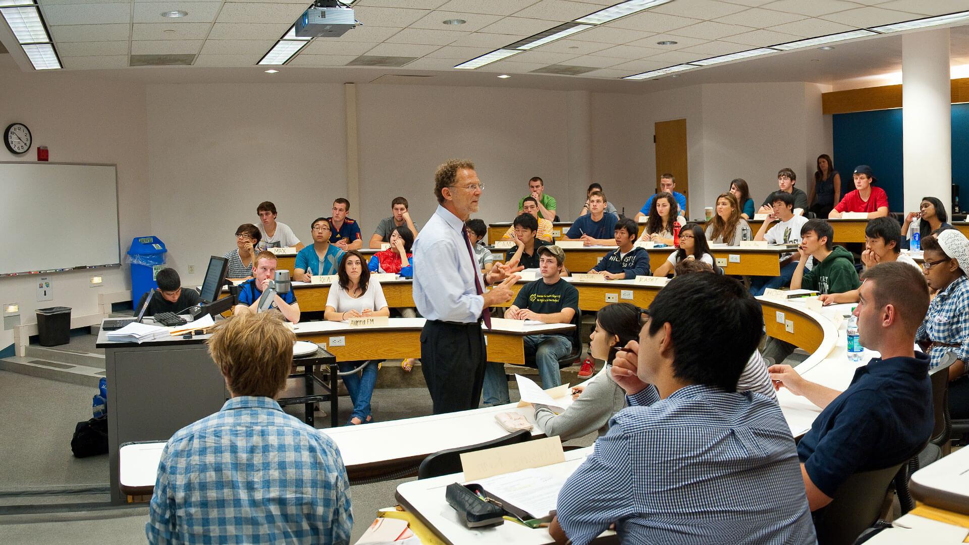 a professor stands in the middle of a lecture hall while students listen intently