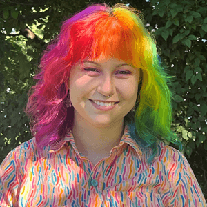 Natalie, smiling happily with multi-colored hair in a shady spot outdoors
