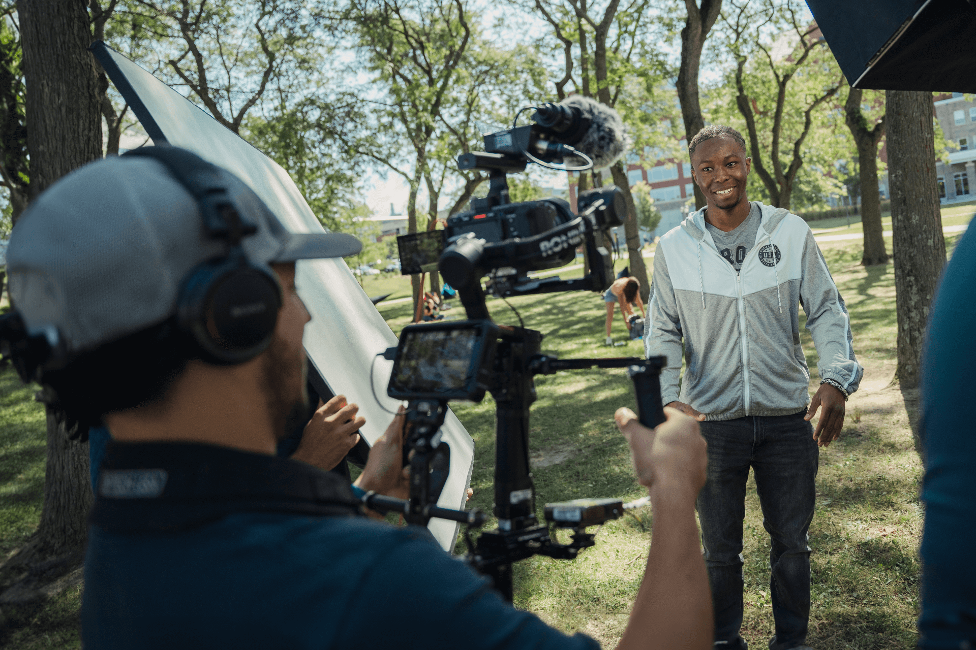 a cameraman holds a camera filming a subject who is smiling outside surrounded by trees