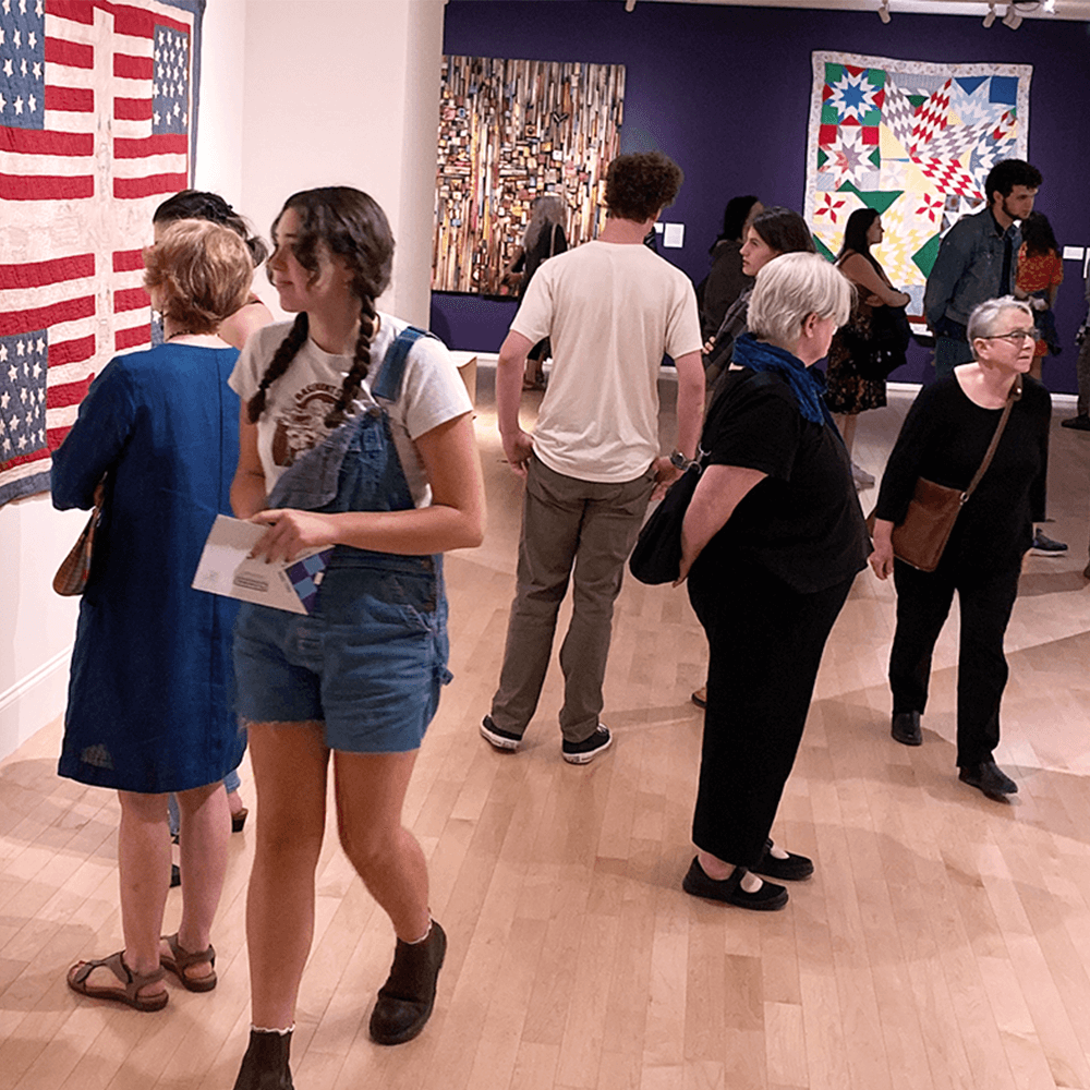 Community members young and old walk through a gallery with many quilts on the wall.