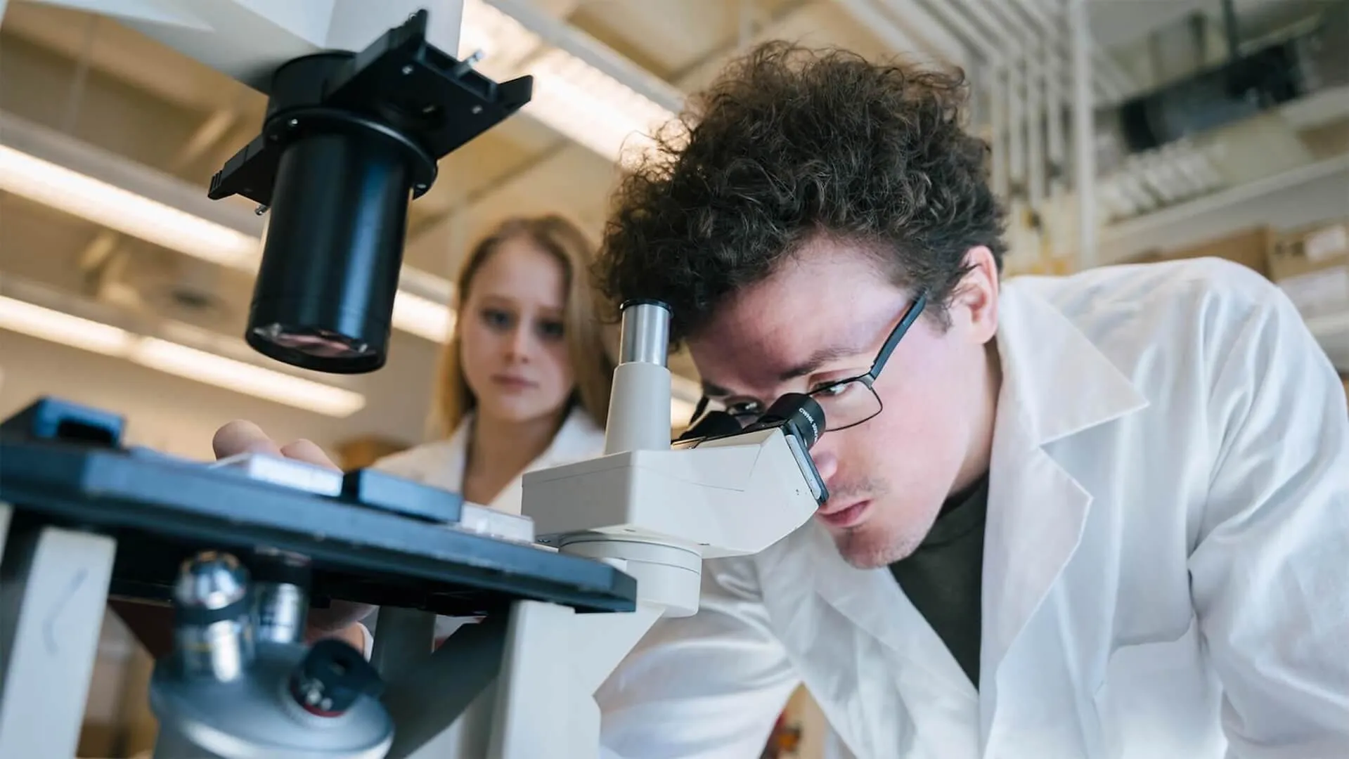 White-coated scientist looks through mictoscope with colleague behind