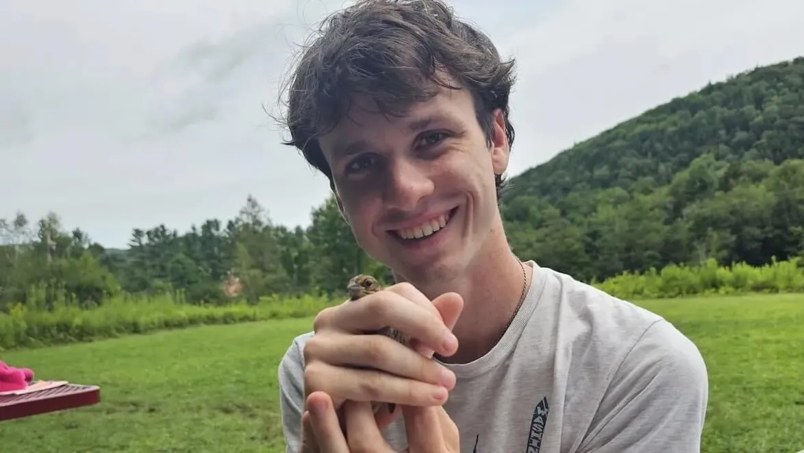 Pierre smiling and holding a small brown bird on a field of green grass with a hill in the background