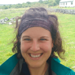 Angie in a headband and rain jacket, smiling broadly, standing in a green field