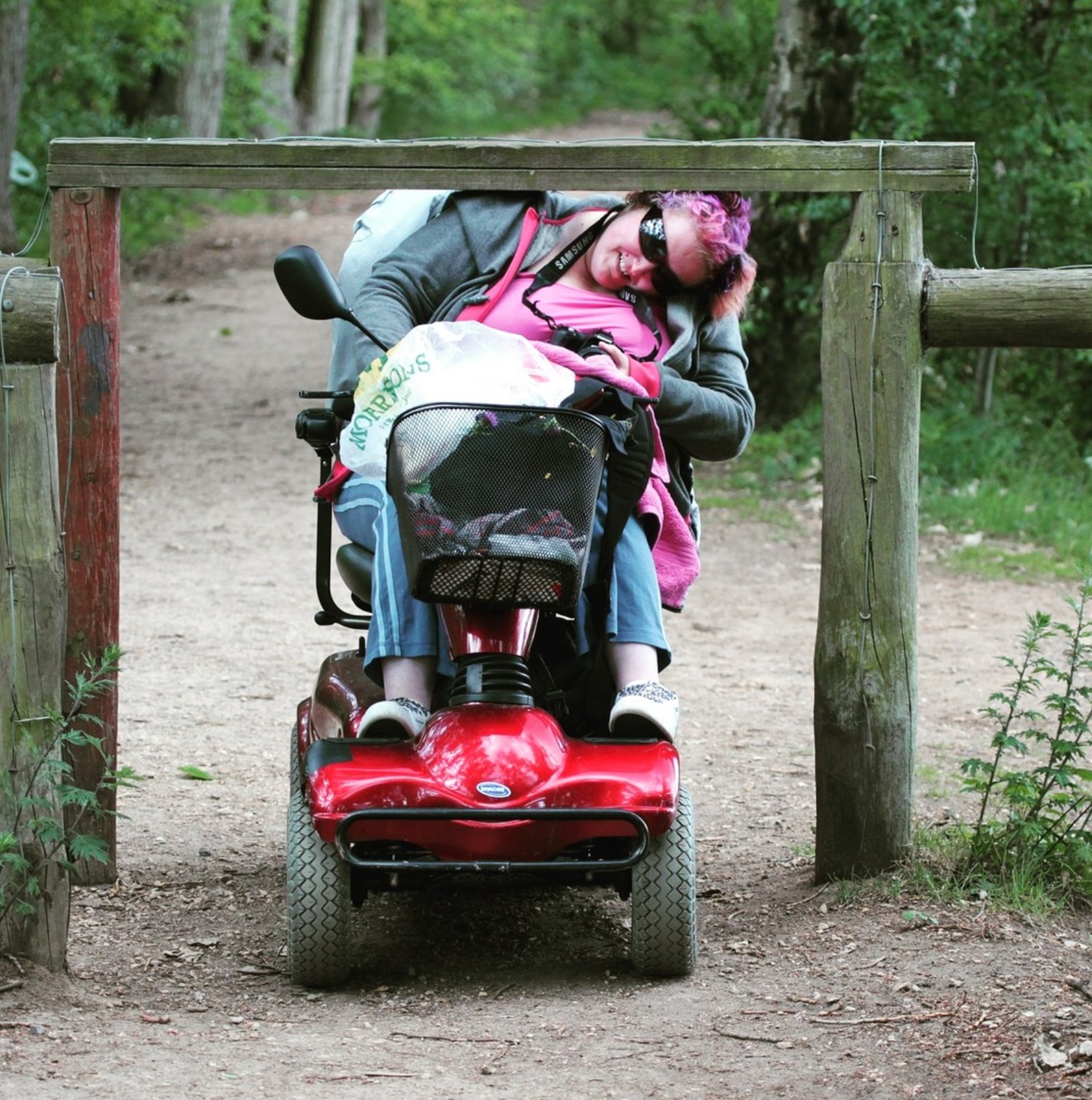 A pink-haired woman in sunglasses and a wheelchair cranes her neck to one side to roll under a wooden crossbeam blocking a dirt path through a forest.