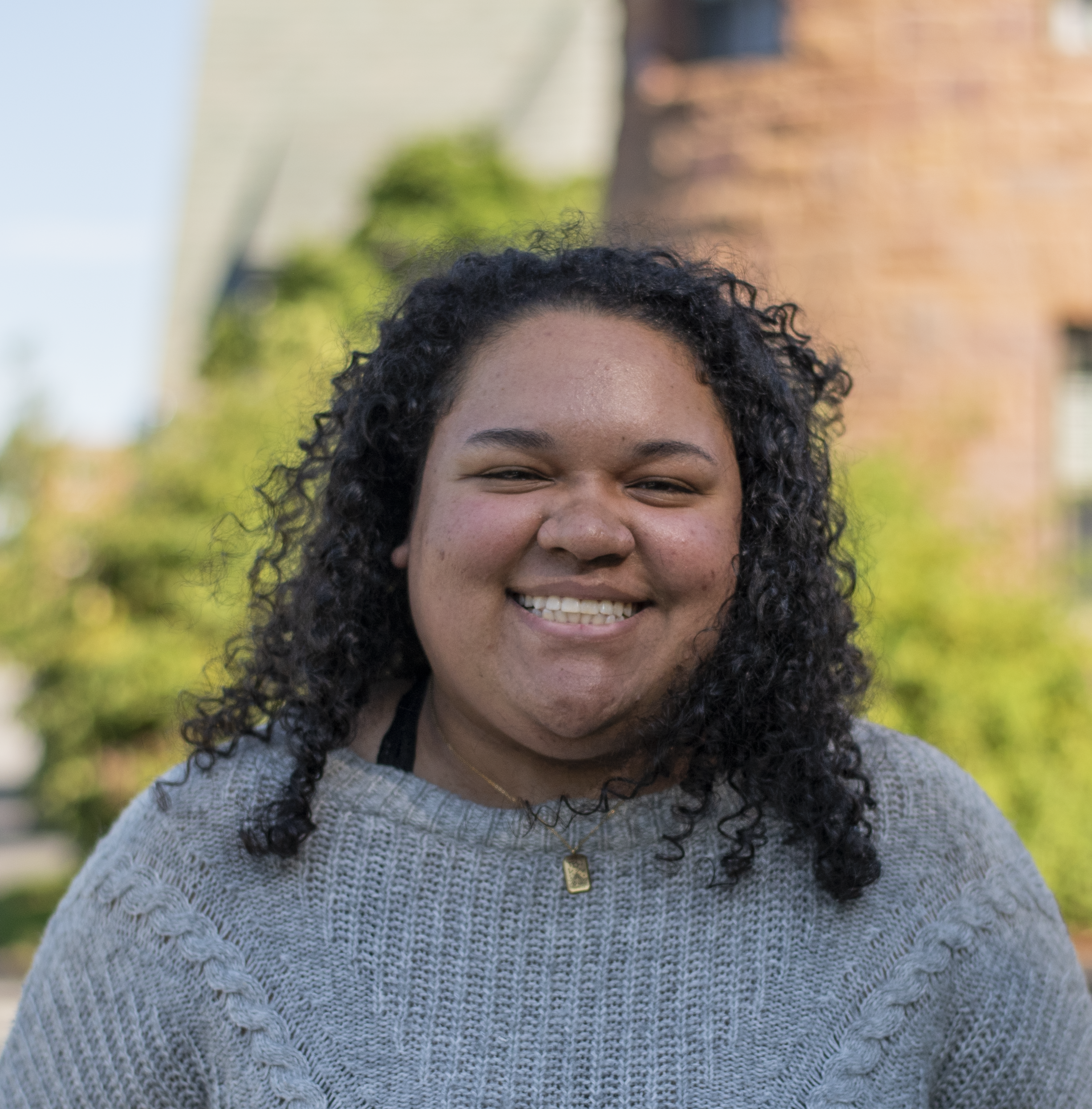 Kay in front of a green leafy background smiling, wearing a grey sweater.